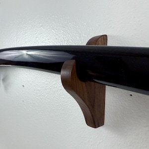 May include: A black katana sword with a dark wood scabbard is mounted on a dark wood wall mount. The sword is secured to the mount with a green cord.