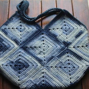 Op de afbeelding: Een gehaakte draagtas in een blauw en grijs kleurenschema. De tas heeft een vierkant patroon en een V-vormige opening aan de bovenkant.