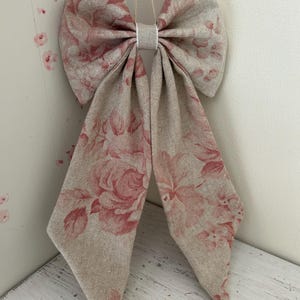 May include: A large decorative bow made from beige fabric, featuring a pink floral design. The bow is tied in the centre and has long, pointed tails. It is suspended against a white wall.