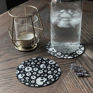 May include: A gold metal candle holder with a glass insert, a glass of water with ice, and two black and white skull-patterned coasters on a wooden surface. One coaster is under the glass, and the other is next to it.