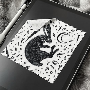 Puede incluir: Un paño de limpieza de microfibra en blanco y negro con una ilustración de un conejo, una luna creciente y estrellas. El paño está diseñado para limpiar pantallas y otras superficies. El diseño tiene un tema celestial.