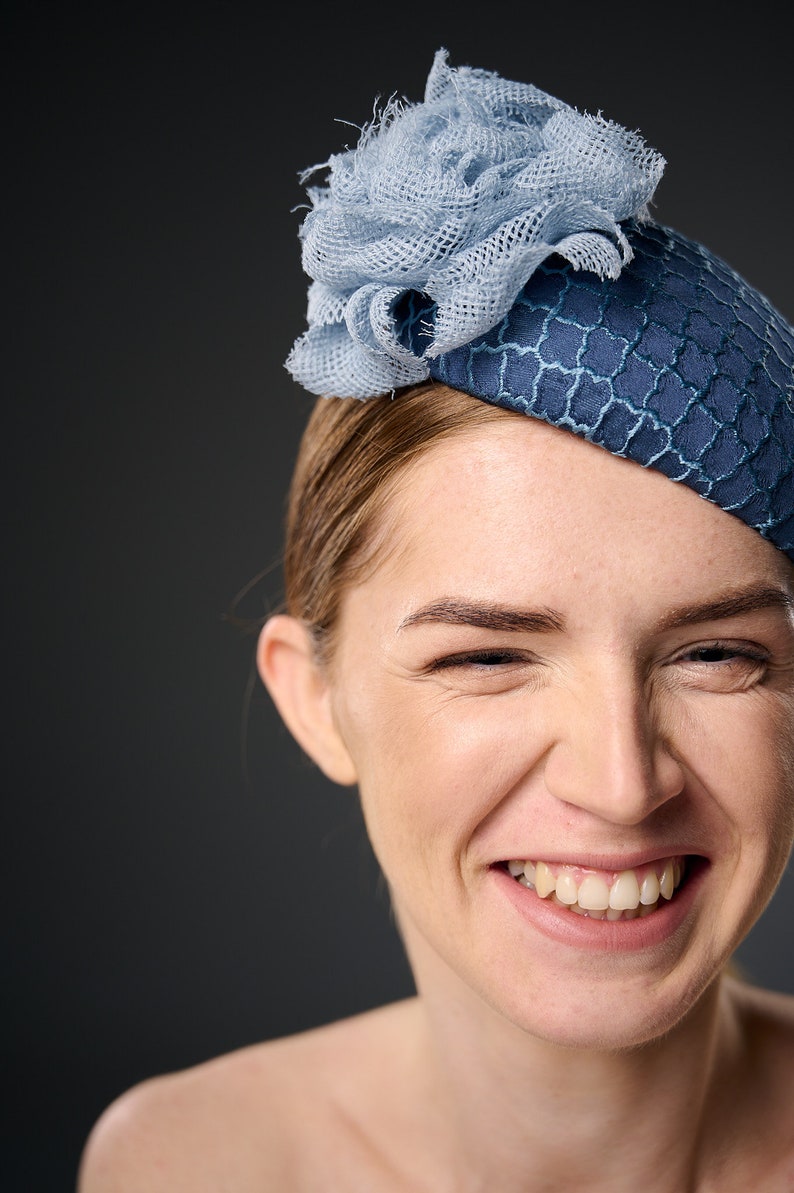 May include: A woman wearing a blue and light blue hat with a flower design. She is smiling and looking at the camera.