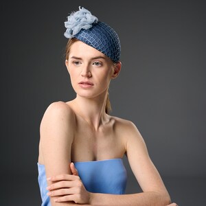 May include: A woman wearing a blue hat with a flower on top. She is sitting at a white table with her arms crossed.