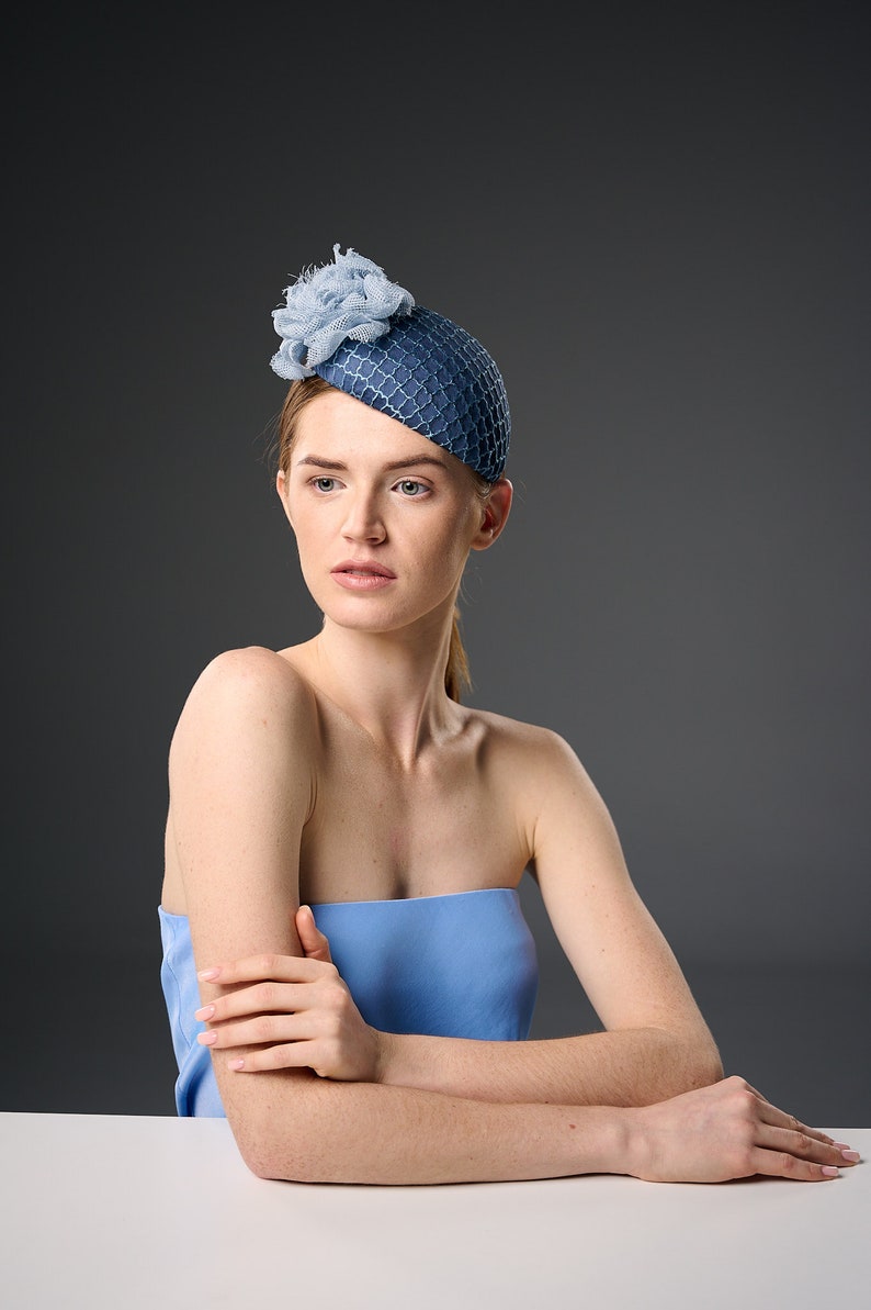 May include: A woman wearing a blue hat with a flower on top. She is sitting at a white table with her arms crossed.