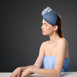 May include: A woman wearing a blue and white patterned hat with a flower accent. She is sitting at a table with her arms resting on the surface.