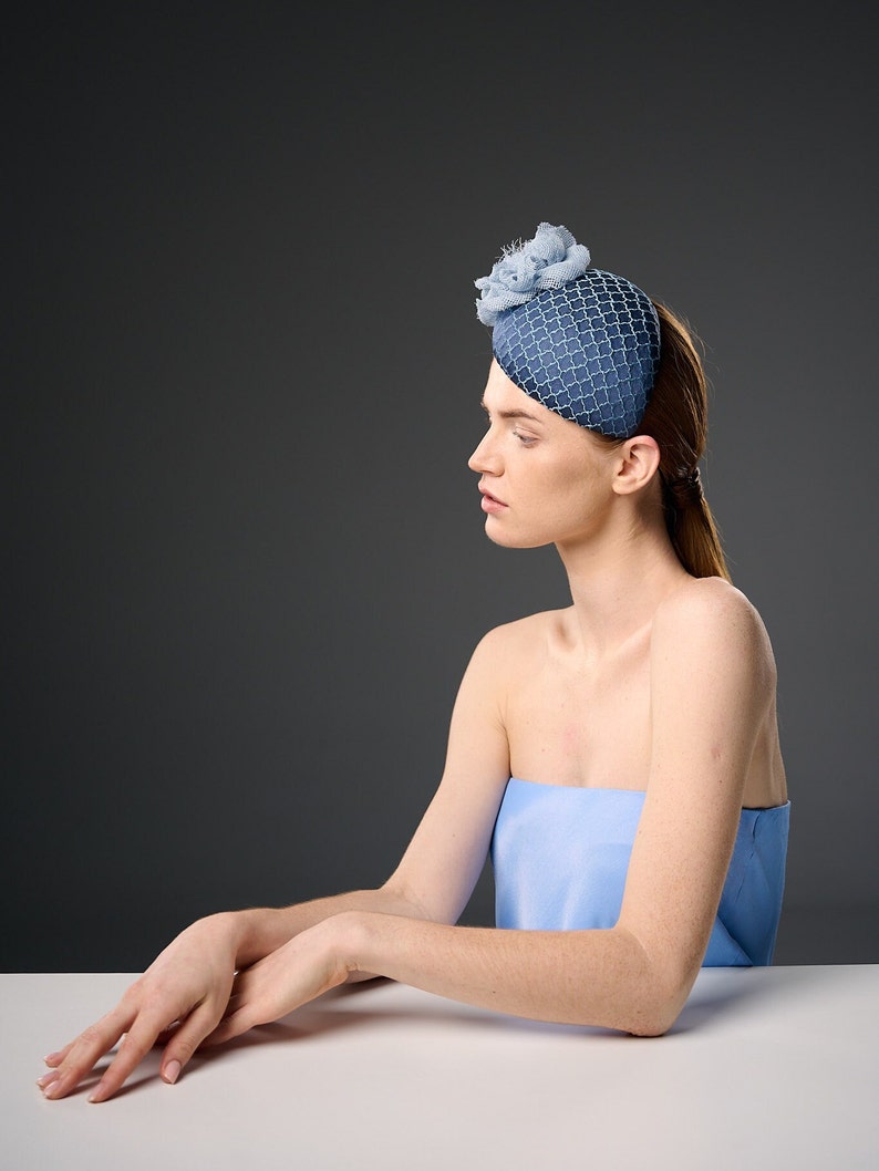 May include: A woman wearing a blue and white patterned hat with a flower accent. She is sitting at a table with her arms resting on the surface.