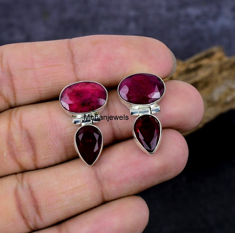 May include: A pair of sterling silver earrings featuring oval-cut ruby gemstones at the top and pear-shaped garnet gemstones at the bottom. The earrings have a polished finish and are set against a dark background.