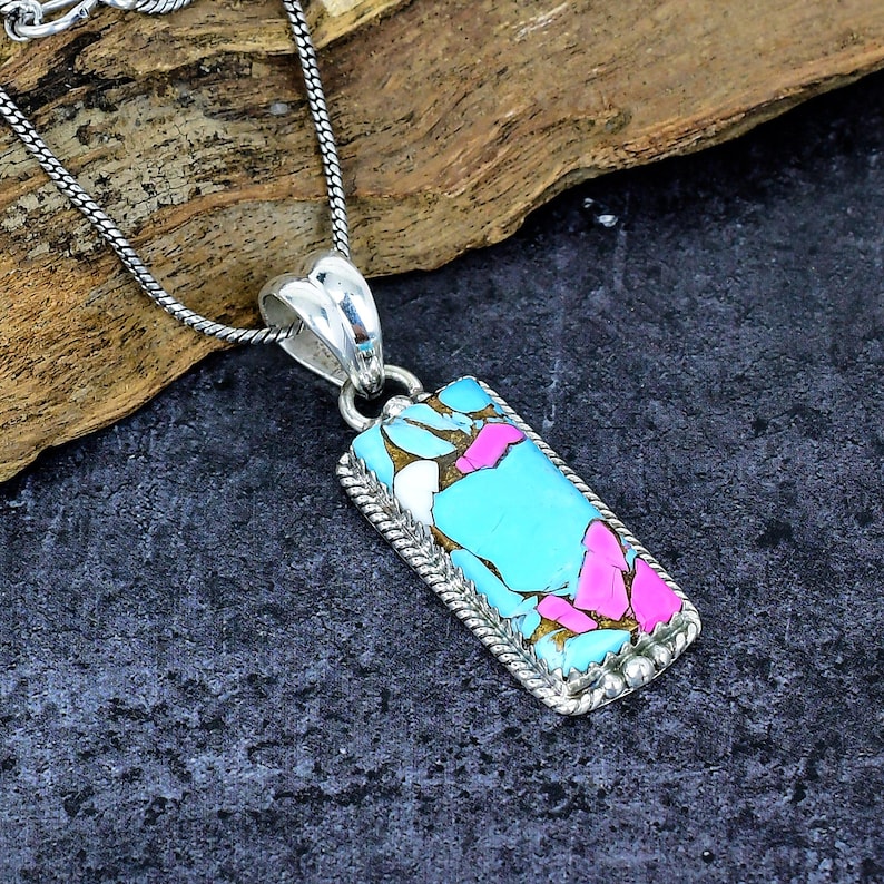 May include: A rectangular pendant necklace with a silver chain. The pendant features a turquoise, pink, white, and gold stone set in a silver frame. The stone has a unique, colorful pattern.