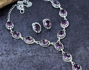 May include: A silver necklace and earring set with oval, deep purple gemstones. The necklace has a pendant design with a series of stones, and the earrings are simple studs. The jewellery is displayed on a dark, textured surface.
