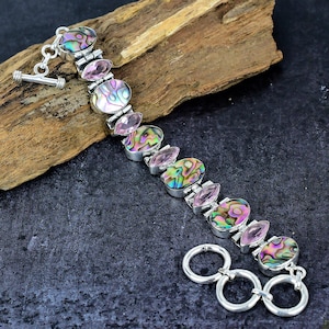 May include: A silver bracelet featuring oval abalone shell and pink gemstone accents. The bracelet has a toggle clasp and is displayed on a piece of wood. The abalone shells have iridescent colors of green, purple, and gold.