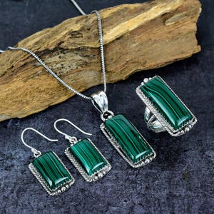 May include: A jewellery set comprising a necklace, ring, and earrings. Each item features a rectangular, green malachite stone set in a silver-coloured metal frame. The earrings are hook-style, and the ring has a wide band.