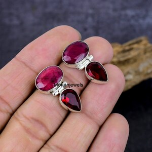 May include: Two pairs of gemstone earrings. Each earring features a large, oval-cut, deep red gemstone paired with a smaller, teardrop-shaped, dark red gemstone, set in a silver metal. The earrings are displayed against a neutral background.