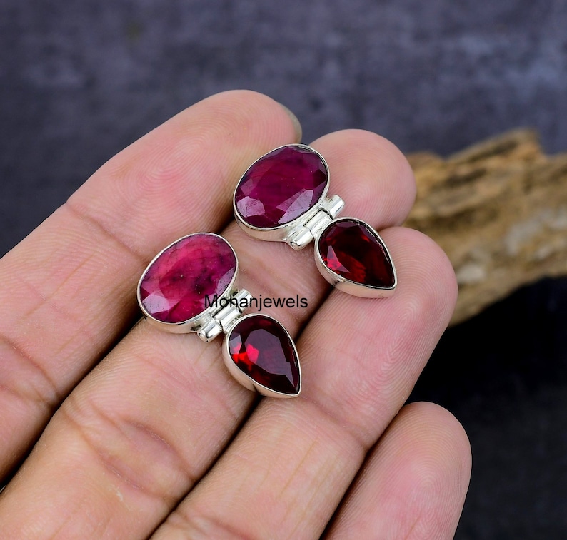 May include: Two pairs of gemstone earrings. Each earring features a large, oval-cut, deep red gemstone paired with a smaller, teardrop-shaped, dark red gemstone, set in a silver metal. The earrings are displayed against a neutral background.