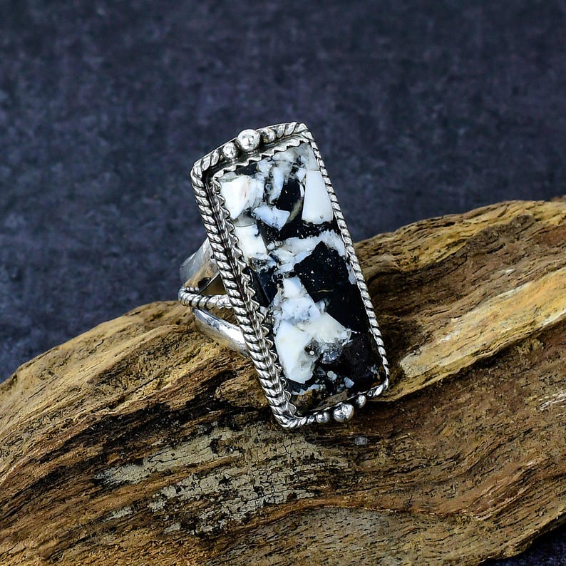 Puede incluir: Anillo de plata con una piedra rectangular con un patr&oacute;n en blanco y negro. La piedra est&aacute; engastada en un marco de plata decorativo con un borde en forma de cuerda. El anillo se muestra sobre un trozo de madera.