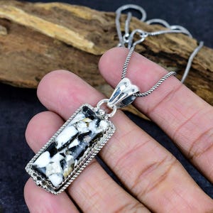 Puede incluir: Collar con colgante rectangular y cadena de plata. El colgante presenta una piedra negra, blanca y marr&oacute;n en un marco plateado con un borde en forma de cuerda. El collar se muestra sobre un fondo de madera.