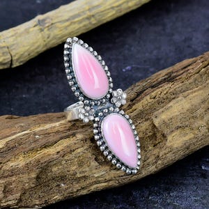 Peut inclure: Une bague en argent ornée de deux pierres roses allongées en forme de goutte, disposées verticalement. La monture métallique est décorée de petits bords perlés et de deux minuscules motifs floraux entre les pierres. L'anneau est lisse.