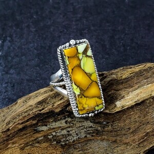 May include: A silver ring with a rectangular, yellow and green stone. The stone has a cracked appearance and is framed by a twisted silver rope design. The ring is set on a piece of wood.