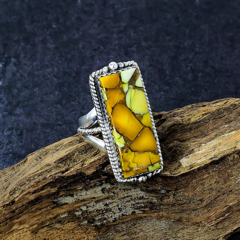 May include: A silver ring with a rectangular, yellow and green stone. The stone has a cracked appearance and is framed by a twisted silver rope design. The ring is set on a piece of wood.