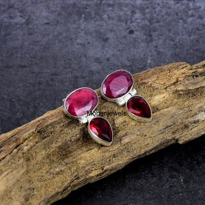 May include: A pair of sterling silver earrings with oval and teardrop-shaped, deep red gemstones. The earrings are set on a wooden surface with a dark background. The gemstones have a faceted cut, reflecting light.
