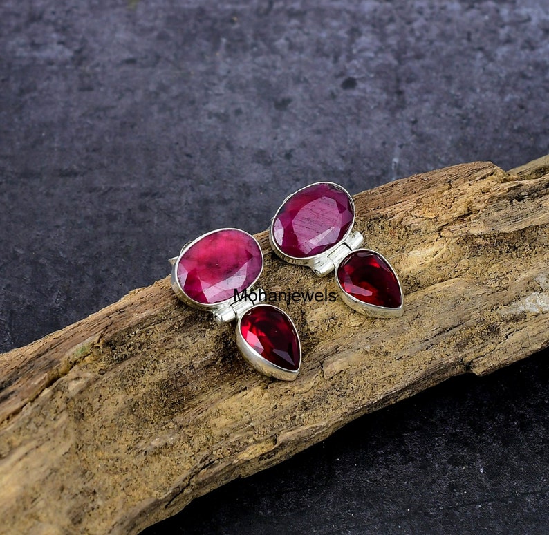 May include: A pair of sterling silver earrings with oval and teardrop-shaped, deep red gemstones. The earrings are set on a wooden surface with a dark background. The gemstones have a faceted cut, reflecting light.