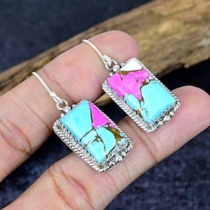May include: A pair of rectangular earrings with a silver-colored frame. The earrings feature a colorful stone with a mix of turquoise, pink, white, and brown hues. The earrings are displayed on a hand.