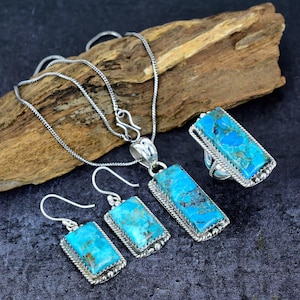 May include: A silver jewelry set featuring turquoise stones. The set includes a pendant necklace, a ring, and a pair of earrings. Each piece has a rectangular turquoise stone with a silver border. The jewelry is displayed on a dark surface with a piece of wood.