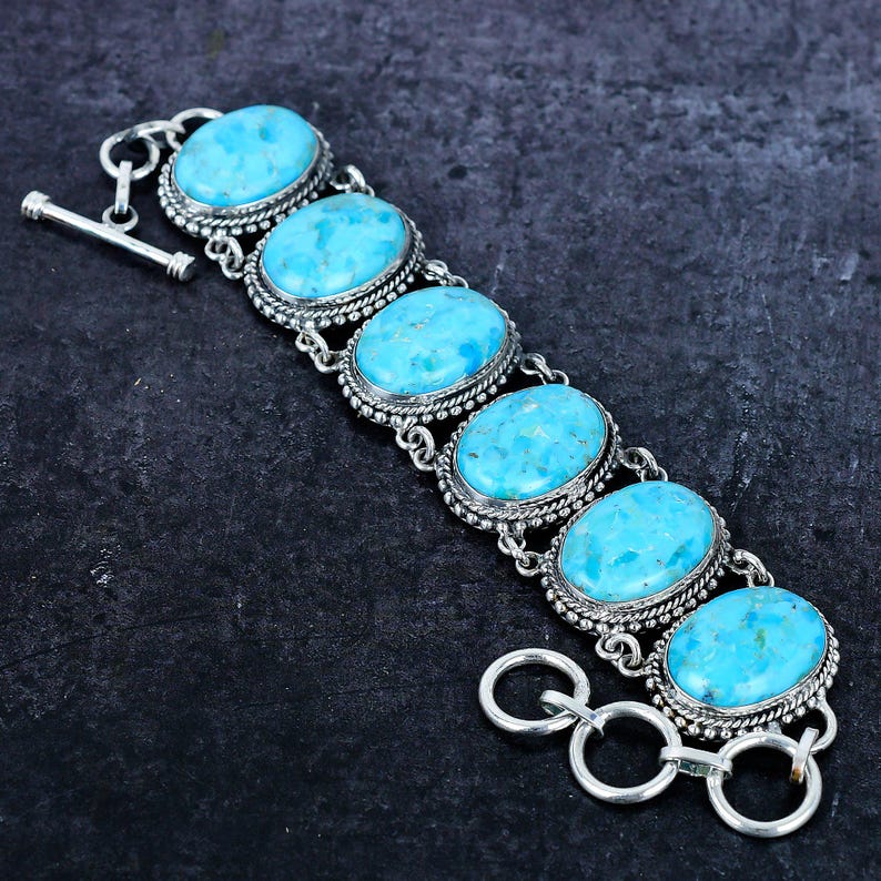 Op de afbeelding: Een zilveren armband met vijf ovale turkoois stenen. Elke steen is gezet in een decoratief zilveren frame met een kralenrand. De armband heeft een tuimelsluiting en wordt getoond tegen een donkere achtergrond.