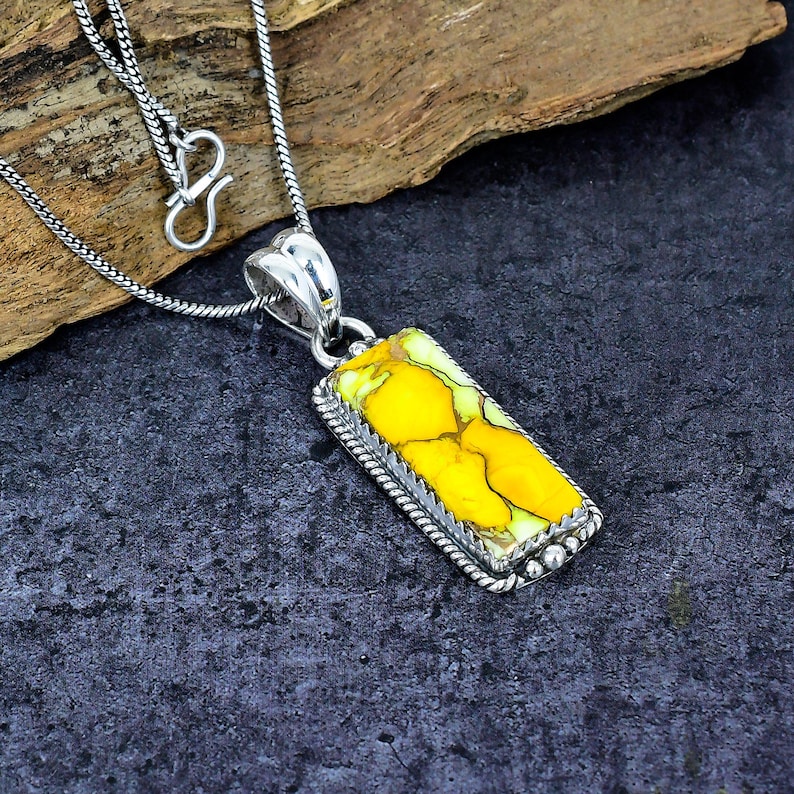 May include: Silver pendant necklace featuring a rectangular yellow stone with a unique pattern. The stone is set in a silver frame with a rope-like border and small bead accents. The necklace has a delicate silver chain.