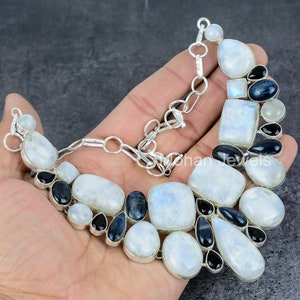 May include: A silver necklace featuring a large, crescent-shaped moonstone pendant and smaller moonstone and black onyx stones. The necklace is displayed on a hand.