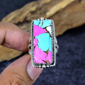 May include: A rectangular silver ring featuring a vibrant, multi-colored stone. The stone is a mosaic of pink, turquoise, white, and brown hues. The ring has a detailed, rope-like border and is set against a dark background.