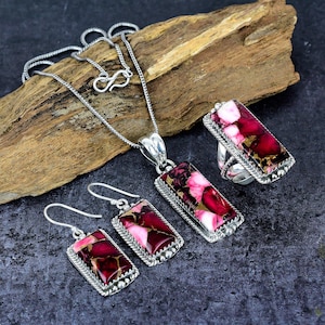 May include: A silver jewelry set featuring a necklace, earrings, and a ring. Each piece has a rectangular, maroon and pink stone with a silver border. The jewelry is displayed on a piece of wood against a dark background.