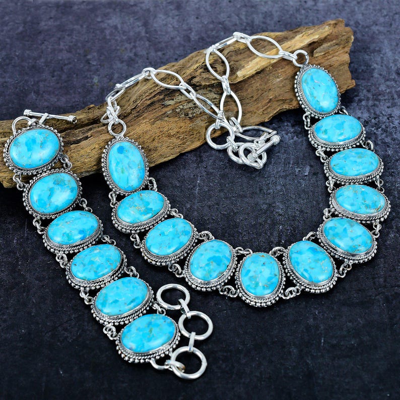 Op de afbeelding: Een zilveren ketting- en armbandset met ovale, lichtblauwe edelstenen. De stenen zijn gezet in een decoratief zilveren frame en verbonden met zilveren kettingen. De sieraden worden gepresenteerd op een stuk hout.