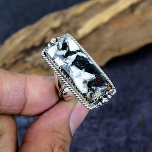 Puede incluir: Anillo rectangular de plata con una piedra grande que muestra un patr&oacute;n veteado de negro, blanco y marr&oacute;n. La piedra est&aacute; engastada en un marco de plata detallado con una textura de cuerda y peque&ntilde;os detalles de cuentas. Un anillo llamativo.