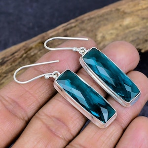 May include: A pair of silver earrings with rectangular, faceted teal-colored gemstones. The gemstones are set in a silver bezel and have a hook closure. The earrings are displayed against a dark background.