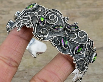 Peut inclure: Bracelet en argent avec un design décoratif orné de plusieurs pierres précieuses irisées vertes et violettes. Le bracelet a une finition texturée et ornée.