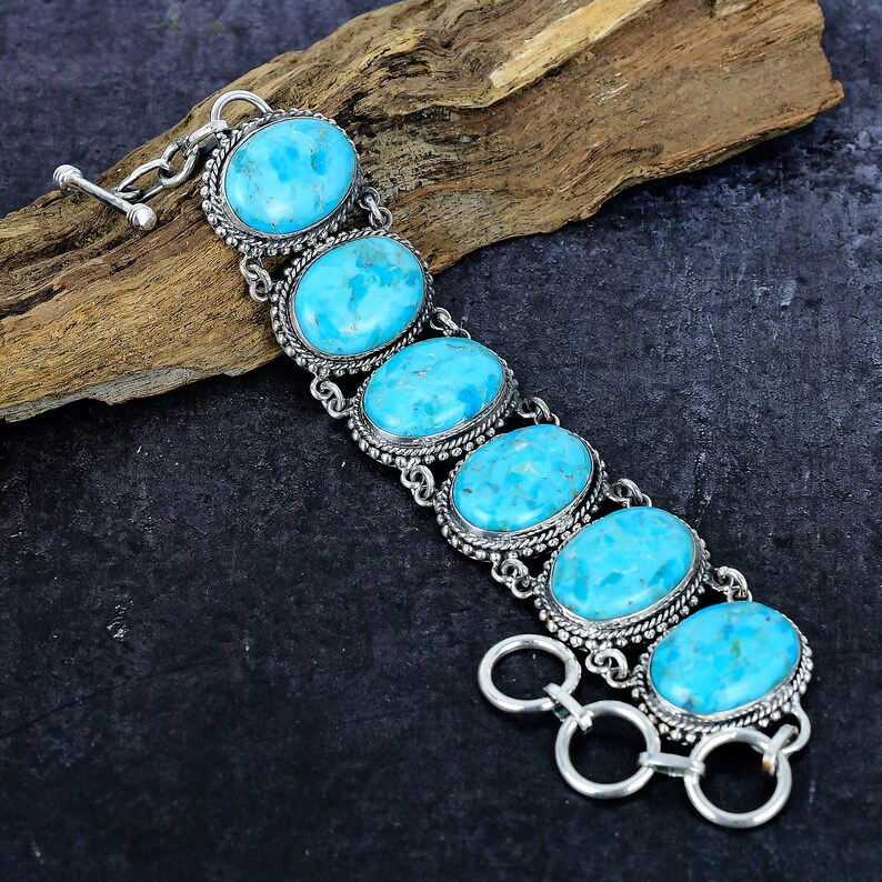 Op de afbeelding: Een zilveren armband met vijf ovale, lichtblauwe edelstenen. Elke steen is gezet in een decoratief zilveren frame met een kralenrand. De armband heeft een tuimelsluiting en wordt gepresenteerd op een donkere ondergrond.