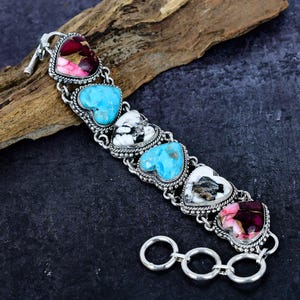 May include: Silver bracelet featuring heart-shaped stones in shades of turquoise, white, and pink. The bracelet has a decorative silver chain and clasp. The stones are set in a silver frame with a rope-like border.