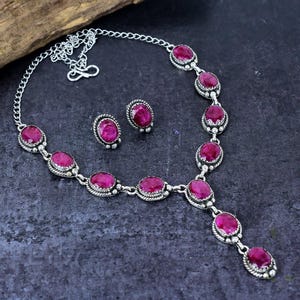 May include: A silver necklace and matching stud earrings set. The jewellery features oval-shaped, deep pink gemstones set in a decorative silver metal. The necklace has a chain and a pendant design. The earrings are simple studs.