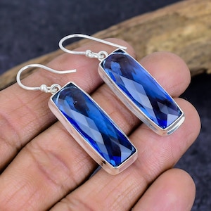 May include: A pair of silver earrings with rectangular, faceted, deep blue gemstones. The gemstones are set in a silver bezel and have a hook closure. The earrings are displayed against a neutral background.
