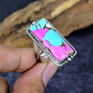 May include: A silver ring with a large, rectangular stone. The stone features a mosaic of pink, turquoise, and white colors. The ring has a textured border and is held by a hand.