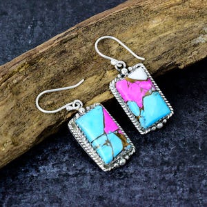 May include: A pair of silver earrings with rectangular turquoise, pink, and white stone inlays. The stones are set in a silver frame with a rope-like border and a decorative silver hook.