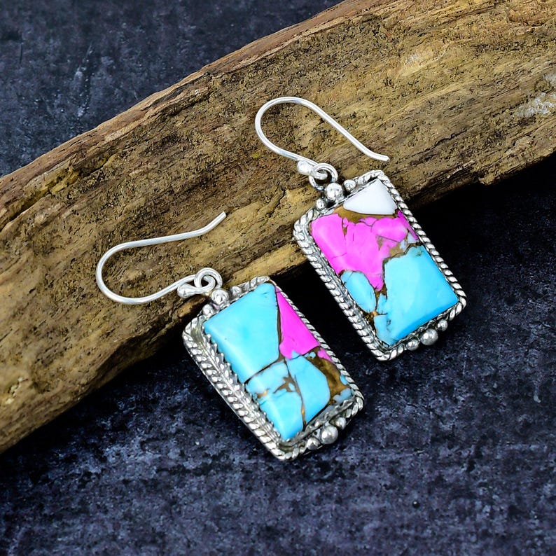 May include: A pair of silver earrings with rectangular turquoise, pink, and white stone inlays. The stones are set in a silver frame with a rope-like border and a decorative silver hook.