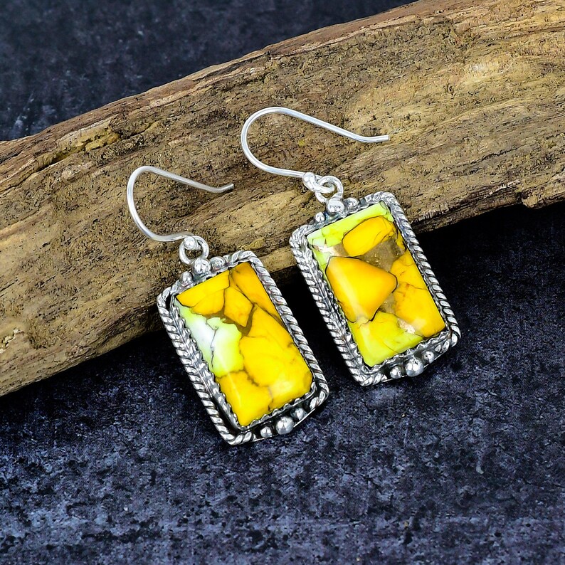 May include: A pair of rectangular earrings with a silver-colored frame. The earrings feature a vibrant yellow and green stone with a textured appearance. The frame has a twisted rope design and small silver beads. The earrings hang from a curved hook.