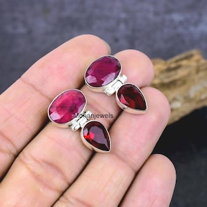 May include: A pair of silver earrings featuring oval-cut ruby gemstones and pear-shaped garnet stones. The earrings are set in a silver metal and are displayed on a hand.