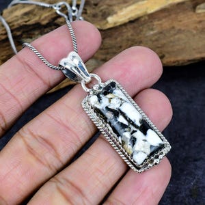Puede incluir: Collar con colgante rectangular. El colgante presenta una piedra blanca, negra y beige engastada en un marco plateado con borde texturizado. La cadena es plateada. Se muestra sobre un fondo oscuro.