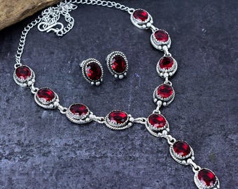 May include: A silver necklace and matching earrings set with oval, deep red gemstones. The necklace has a chain design with multiple stones, and the earrings mirror the necklace stones. The jewellery is displayed on a dark, textured surface.
