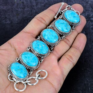 Op de afbeelding: Een zilveren armband met zes ovale turkooizen stenen. Elke steen is gezet in een decoratief zilveren frame met een kralenrand. De armband heeft een tuimelsluiting en wordt op een hand getoond.