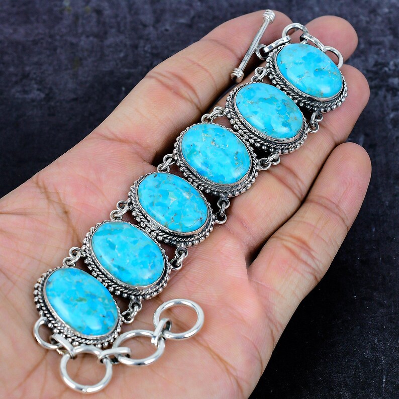 Op de afbeelding: Een zilveren armband met zes ovale turkooizen stenen. Elke steen is gezet in een decoratief zilveren frame met een kralenrand. De armband heeft een tuimelsluiting en wordt op een hand getoond.