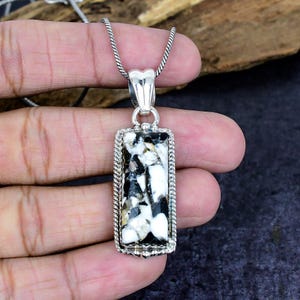 Puede incluir: Un colgante rectangular con una piedra negra y blanca engastada en un marco plateado, suspendido de una cadena plateada. La piedra tiene un aspecto veteado con una mezcla de colores negro, blanco y gris. El marco del colgante tiene un borde texturizado detallado.