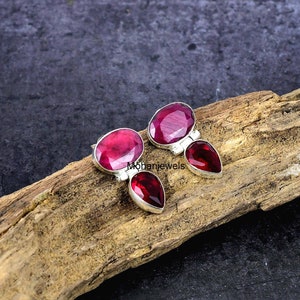 May include: A pair of silver earrings with oval and pear-shaped red gemstones. The earrings are displayed on a piece of wood. The brand name "Mohanjewels" is visible in the image.
